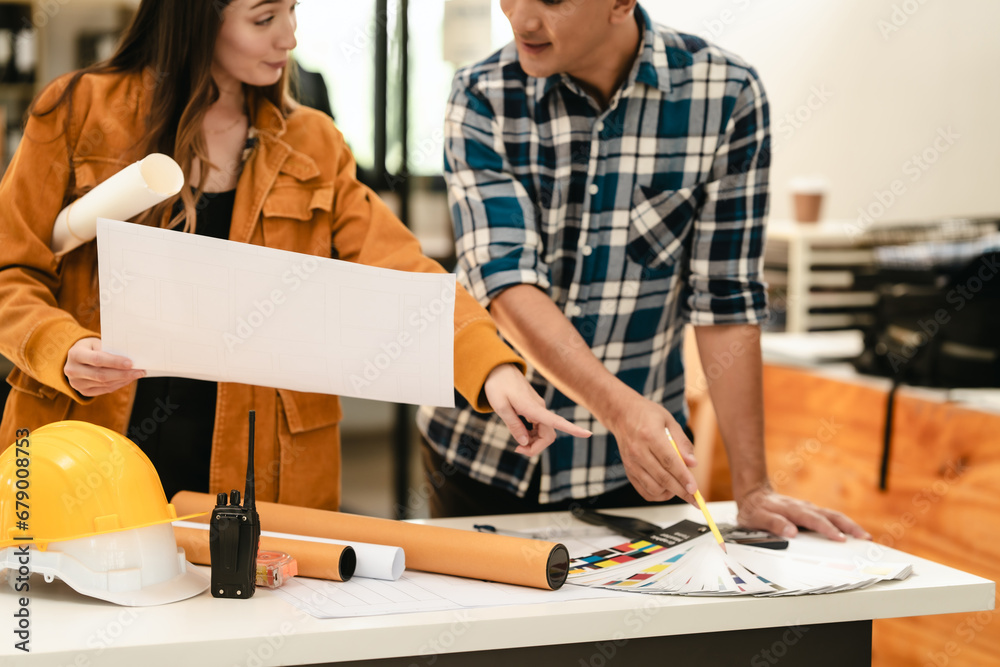 Asian man and Caucasian woman, both engineers, examining set of blueprints closely during team meeting, with house model on the table. Asian man and Caucasian woman, both engineers, examining set of blueprints closely during team meeting, with house model on the table.