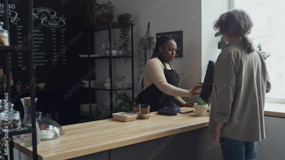 Medium long shot of female customer with grey hair paying for her order with smartphone through terminal with barista standing behind counter in coffee shop Medium long shot of female customer with grey hair paying for her order with smartphone through terminal with barista standing behind counter in coffee shop