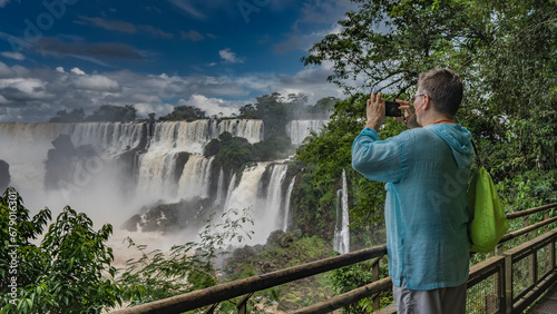 A man stands on the observation deck at the railing, taking pictures of the waterfall on his smartphone. Backpack on the shoulder. Streams of water break from the ledges of the rocks. Spray, fog. photo