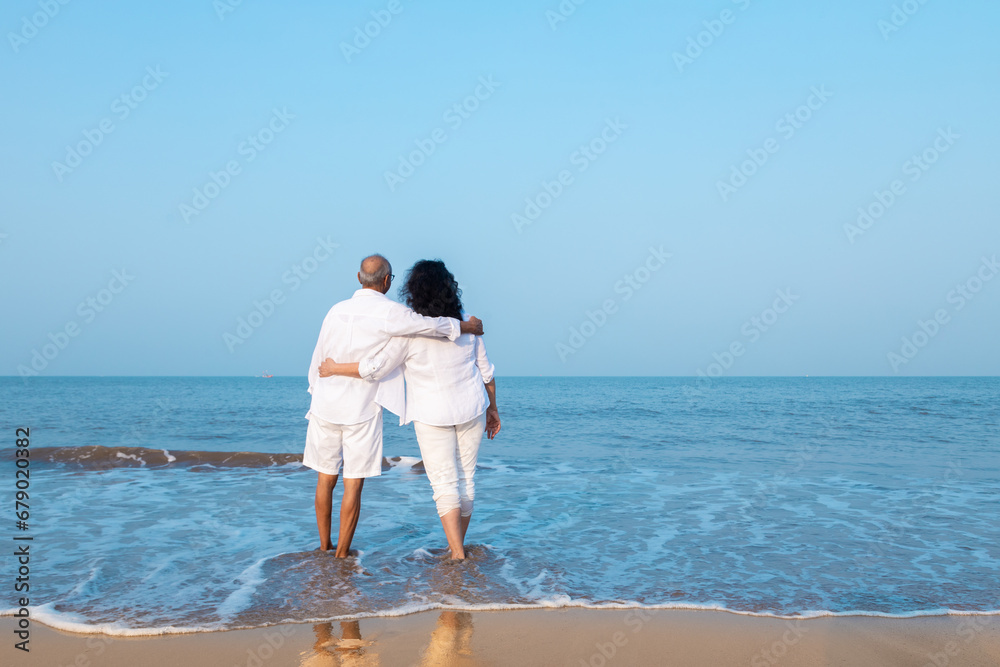 Happy senior indian couple wearing white cloths enjoying summer vacation, holiday at beach looking at sea. Copy space