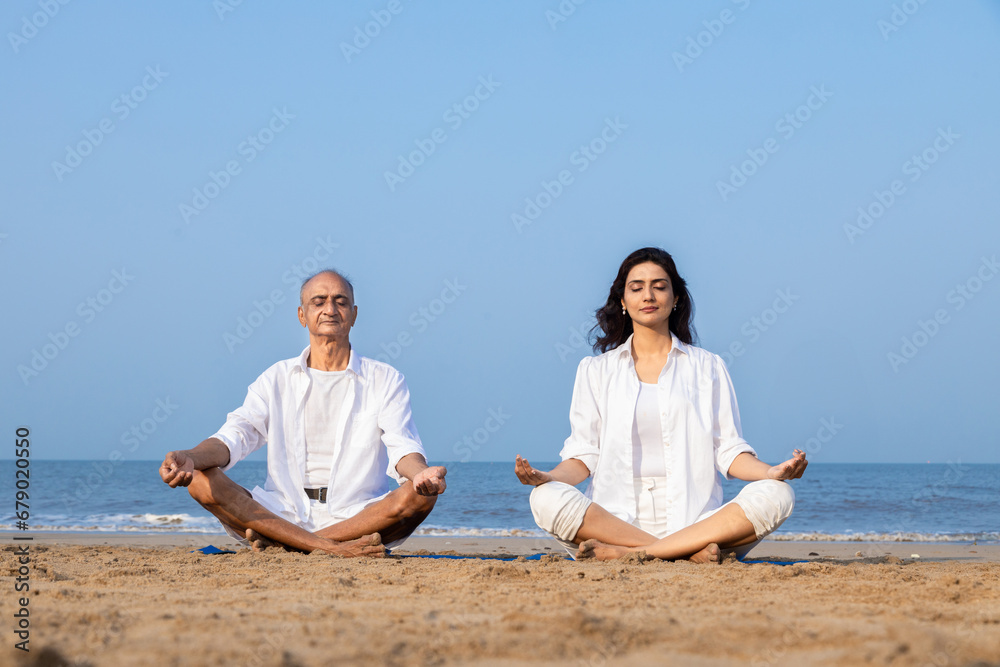 Senior indian father and daughter sitting on fitness mat in lotus position meditating together at beach. Healthy family Concept. Senior indian father and daughter sitting on fitness mat in lotus position meditating together at beach. Healthy family Concept.