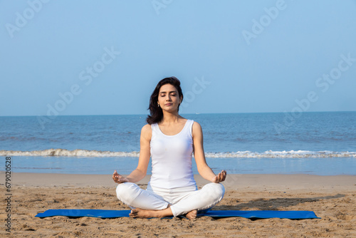 Beautiful indian woman wearing white cloths sitting on fitness mat in lotus position meditating at beach. Healthy Life Concept.Copy space.