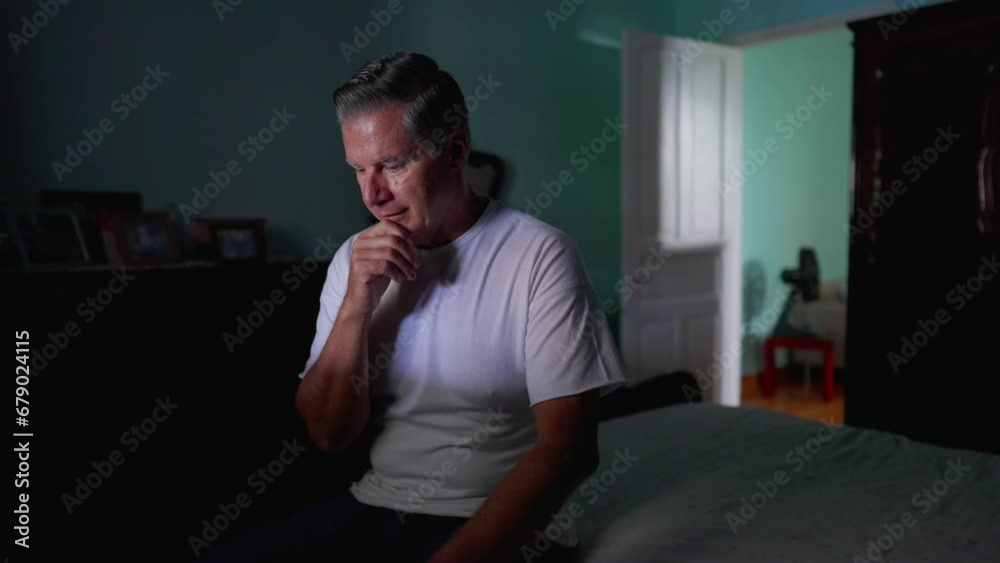 Thoughtful older man sitting by bedside with pensive expression. Domestic lifestyle of middle-aged person pondering solution to problem Thoughtful older man sitting by bedside with pensive expression. Domestic lifestyle of middle-aged person pondering solution to problem