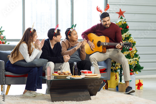 Asian Indian multinational cheerful male female friends celebrating Christmas Eve and Happy New Year wearing reindeer horn headband playing guitar in living room full decoration with Xmas tree