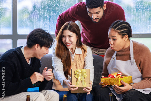 Millennial Asian surprised excited woman model holding opening wrapped present gift box with cheerful male female friends looking cheering in living room celebrating Christmas Eve and New Year