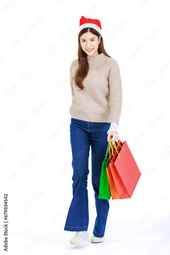 Isolated cutout studio full body shot of Asian cheerful happy beautiful female model wears casual outfit with red Santa Claus hat standing smiling holding shopping bags posing on white background Isolated cutout studio full body shot of Asian cheerful happy beautiful female model wears casual outfit with red Santa Claus hat standing smiling holding shopping bags posing on white background