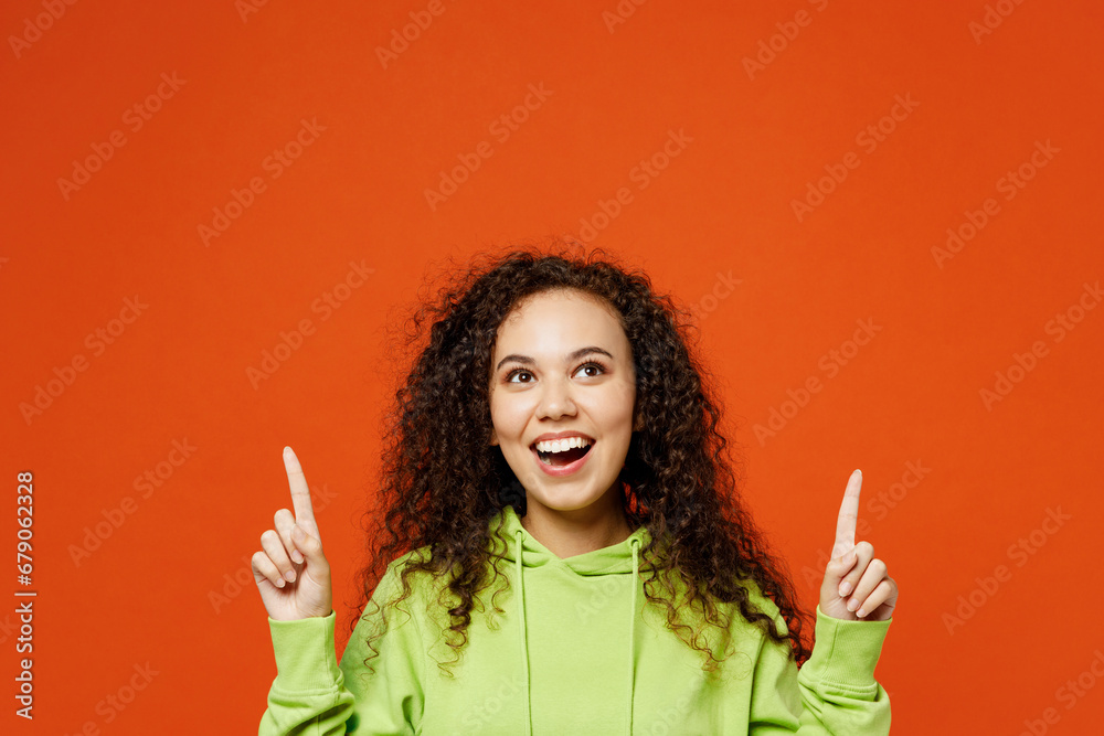 Surprised fun amazed young woman of African American ethnicity wears green hoody casual clothes point index finger overhead on area mockup isolated on plain red orange background. Lifestyle concept. Surprised fun amazed young woman of African American ethnicity wears green hoody casual clothes point index finger overhead on area mockup isolated on plain red orange background. Lifestyle concept.