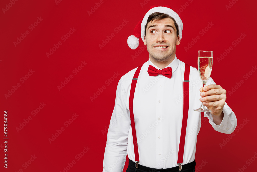 Naklejka premium Merry young smiling man wears white shirt Santa hat posing hold glass with champagne look aside on area isolated on plain red background studio. Happy New Year Christmas celebration holiday concept.