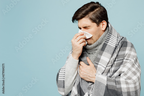 Quadro em tela Young sad ill sick man wrapped in gray plaid sneezing use paper napkin for runny nose isolated on plain blue background studio