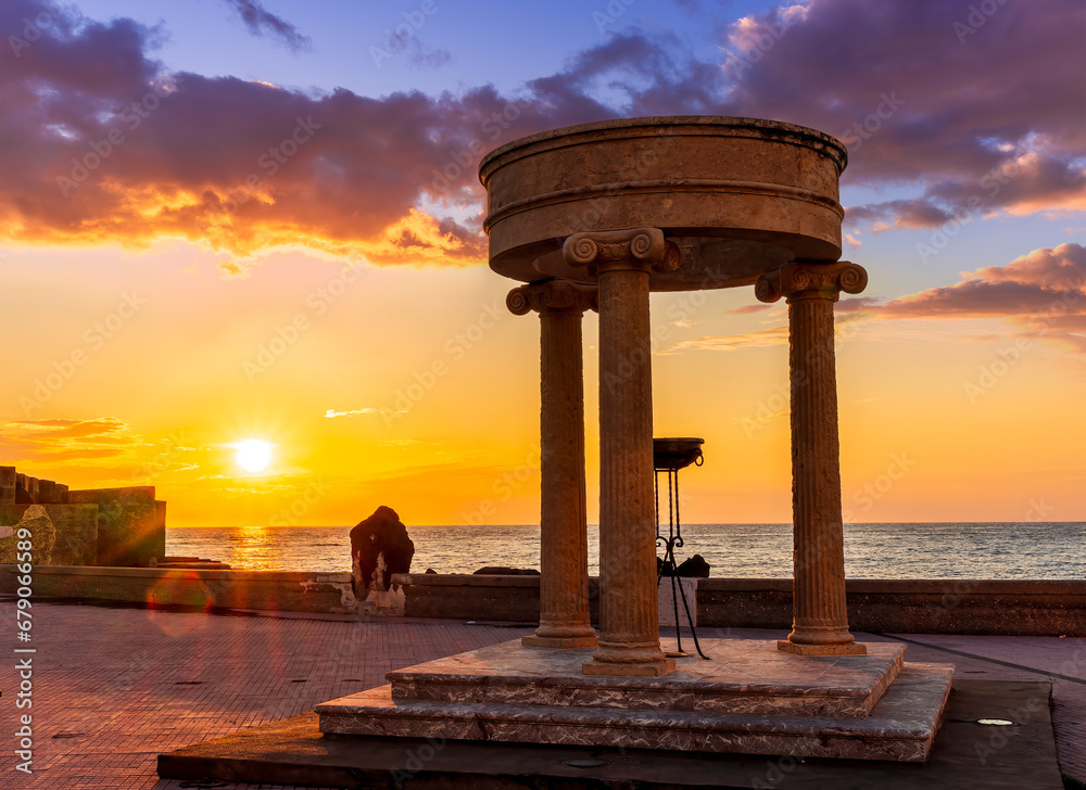 beautiful stone monument with three columns in ancient history greek style with see embarkment and amazing cloudy sunset on background of landscape