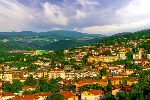 Wallpaper Mural panoramic landscape view of green summer city with yellow houses, green forest and mountains above and amazing hills and cloudy sky on background Torontodigital.ca