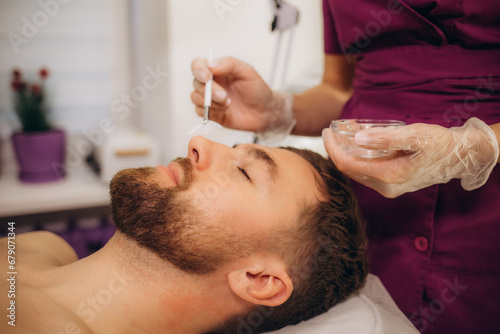 Cosmetologist applying cosmetic mask on man's face in spa salon. Handsome bearded man getting facial skin care treatment.