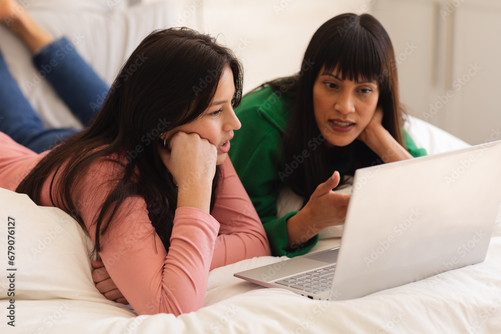 Focused biracial mother and teenage daughter on bed and using laptop at home, copy space Focused biracial mother and teenage daughter on bed and using laptop at home, copy space
