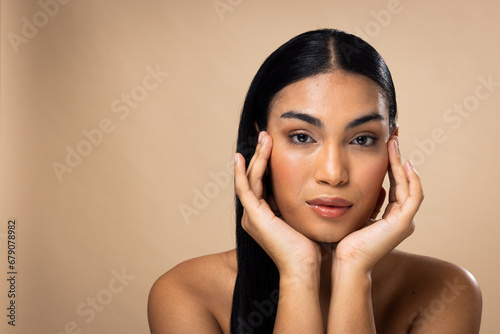 Portrait of biracial woman with dark hair, hands to face and natural make up on brown background photo
