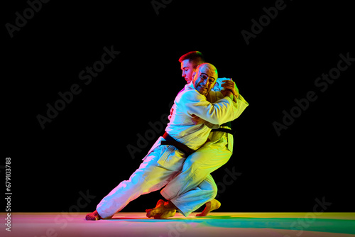 Two young professional karate men fighting, performing technical skill in neon light isolated black background. Copy space for ad, text