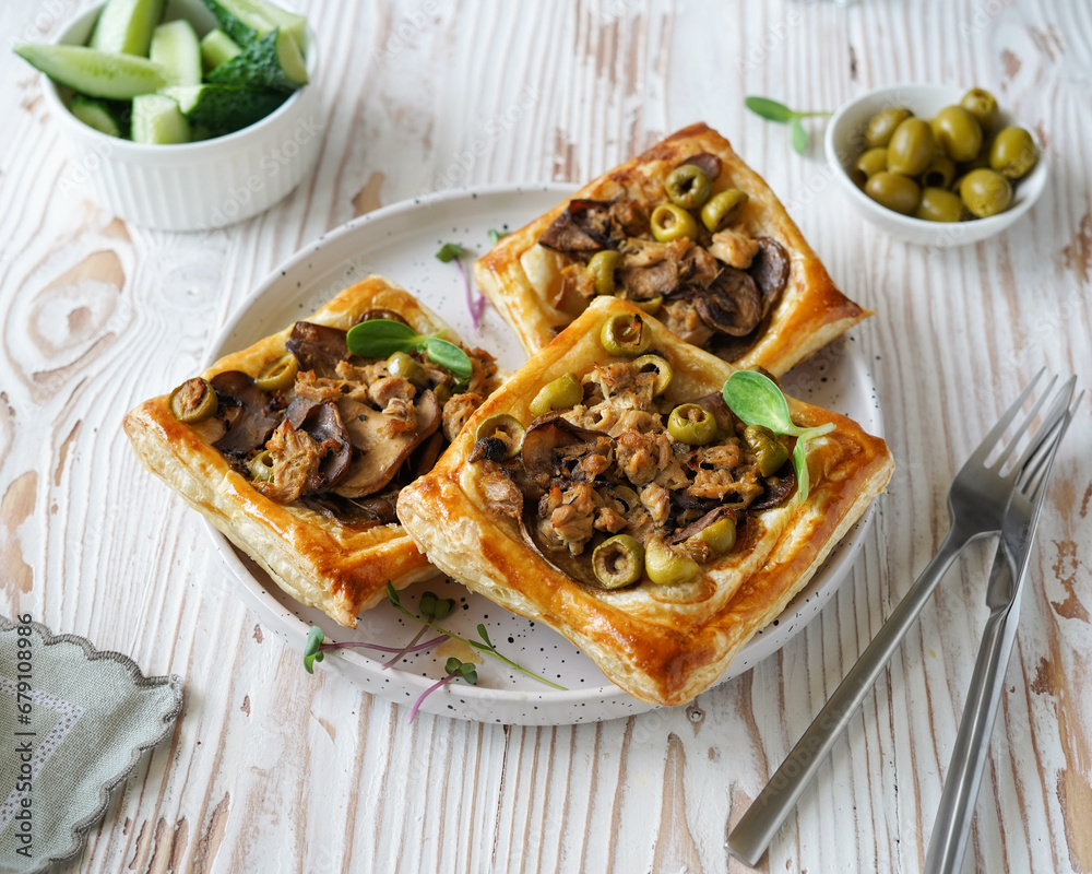 pastry with mushroom and olive filling on puff pastry, white ceramic plate, side bowl with olives, light wooden background, close-up