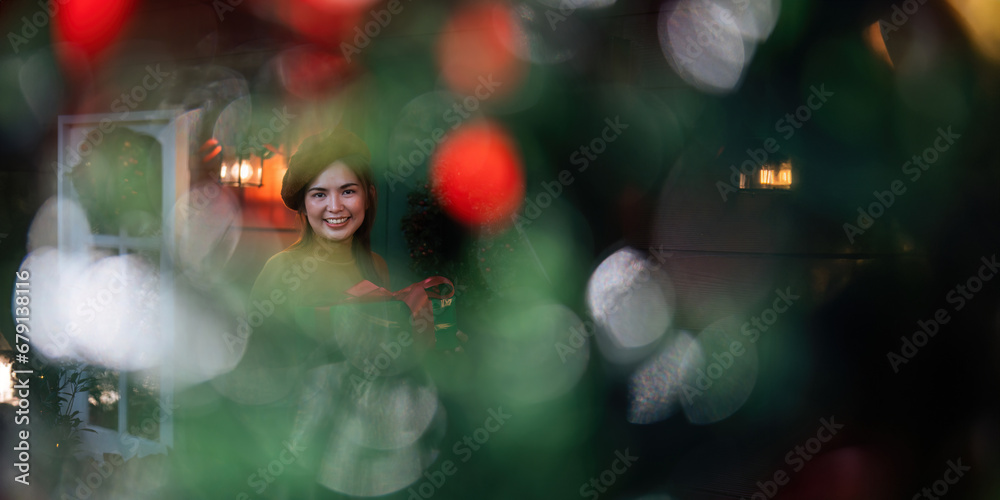 Happy woman wearing Santa hat holding of gift box. Positive emotional Santa girl. with a beautifully decorated Christmas tree serving as the background. festive Xmas concept