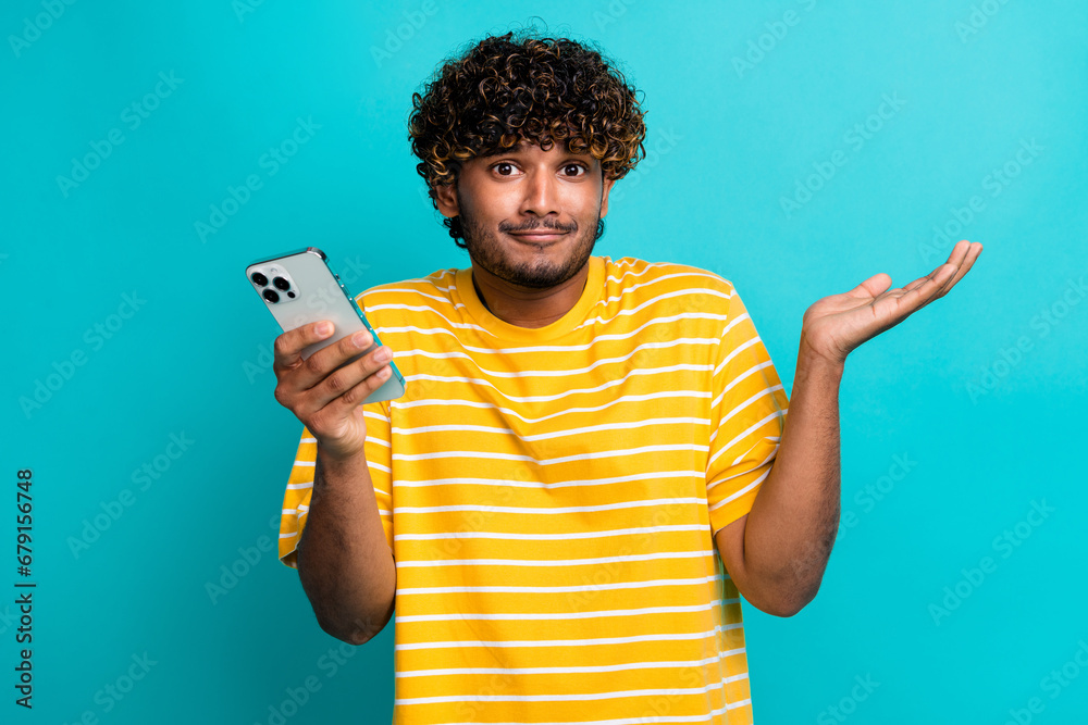 Photo portrait of attractive young man gadget shrug shoulders wear trendy striped yellow clothes isolated on aquamarine color background Photo portrait of attractive young man gadget shrug shoulders wear trendy striped yellow clothes isolated on aquamarine color background