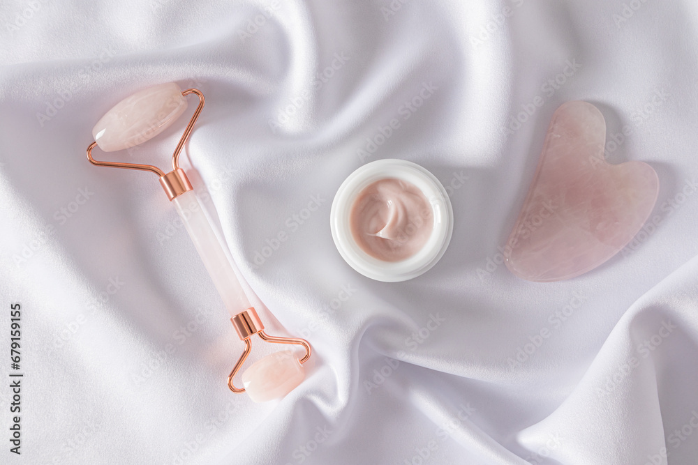 A set of cosmetics for massage and skin care of the face and neck on a soft white satin. quartz roller, gua sha scraper, cream jar. Top view. A set of cosmetics for massage and skin care of the face and neck on a soft white satin. quartz roller, gua sha scraper, cream jar. Top view.