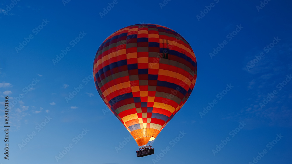 Naklejka premium A colorful balloon in the clear dark sky at dawn, with fire burning inside, and people in the basket expressing joy.