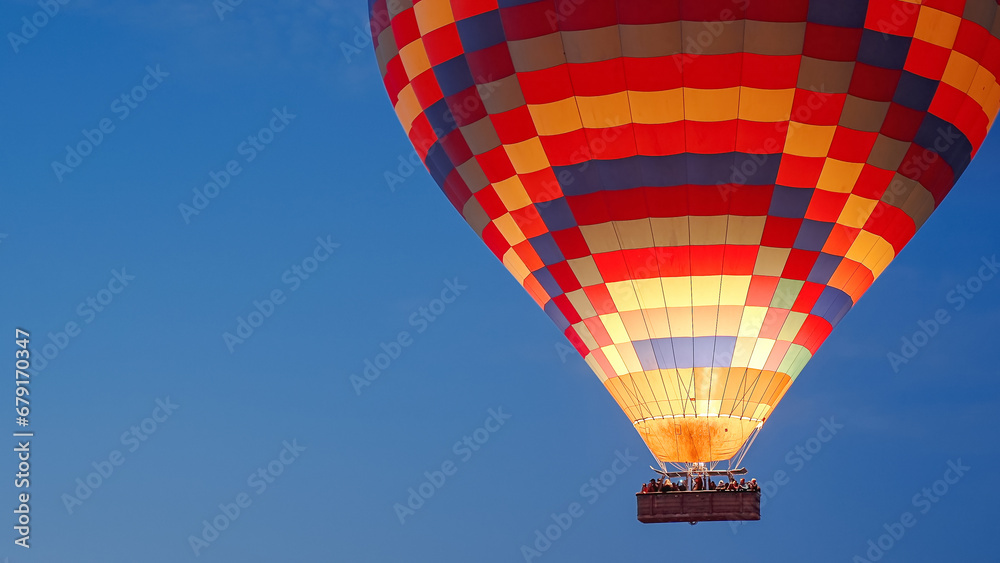 Fototapeta premium A multicolored hot air balloon with people in the basket, against the dark sky at dawn. Fire burns inside.