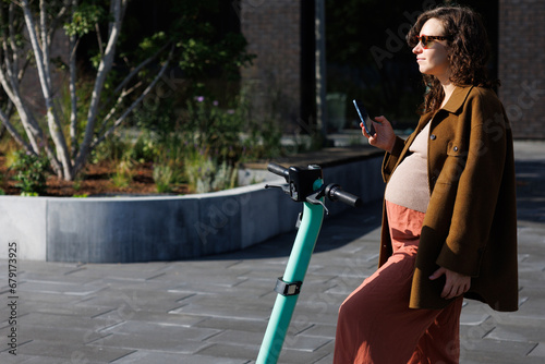 Pregnant woman in daly city life. Female using mobile phone standing next to a electric scooter. Green and sustainable urban city photo