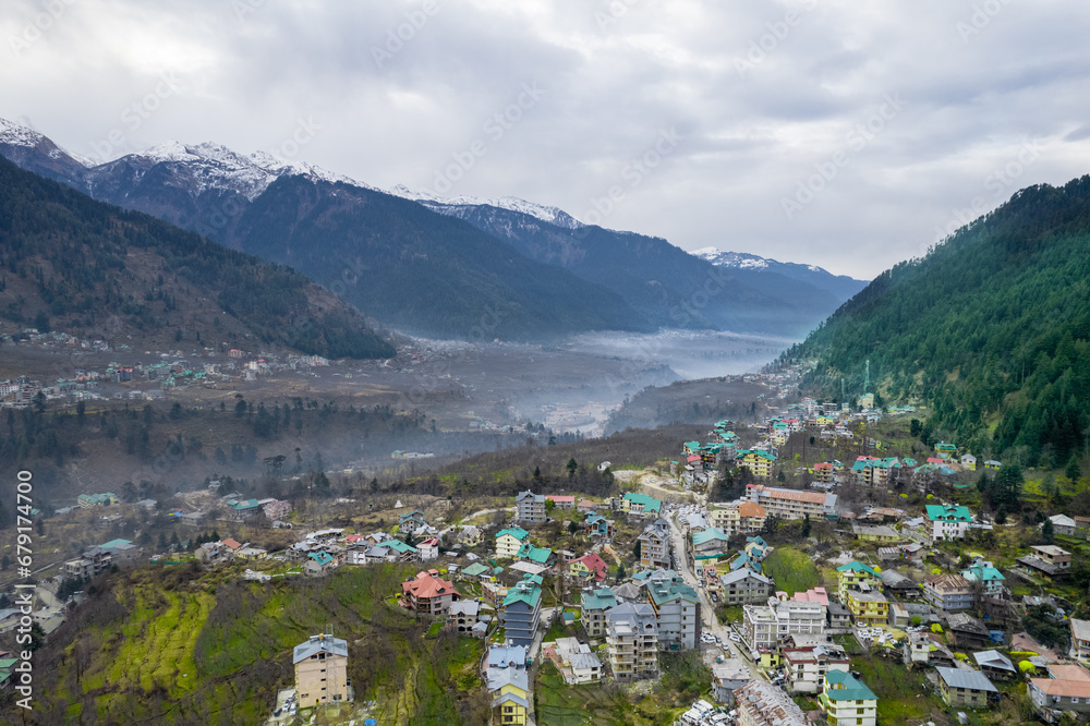 aerial drone shot showing manali kullu town in valley with fog covered himalaya mountains nearby and green fields in between