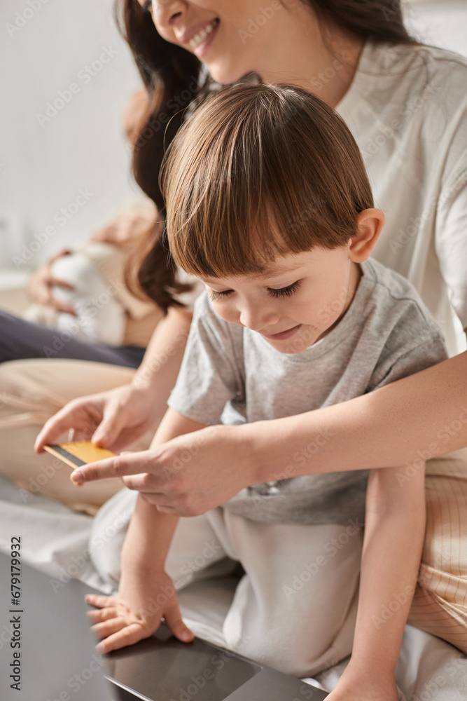 vertical focused shot of little boy looking at laptop next to his blurred mother holding credit card vertical focused shot of little boy looking at laptop next to his blurred mother holding credit card