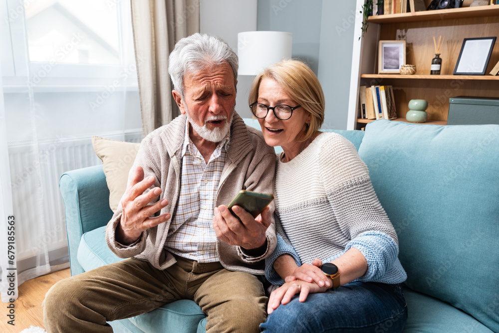 Happy senior old couple holding smartphone looking at cellphone screen laughing relaxing sit on sofa together, smiling elder mature grandparents family embracing having fun with mobile phone at home