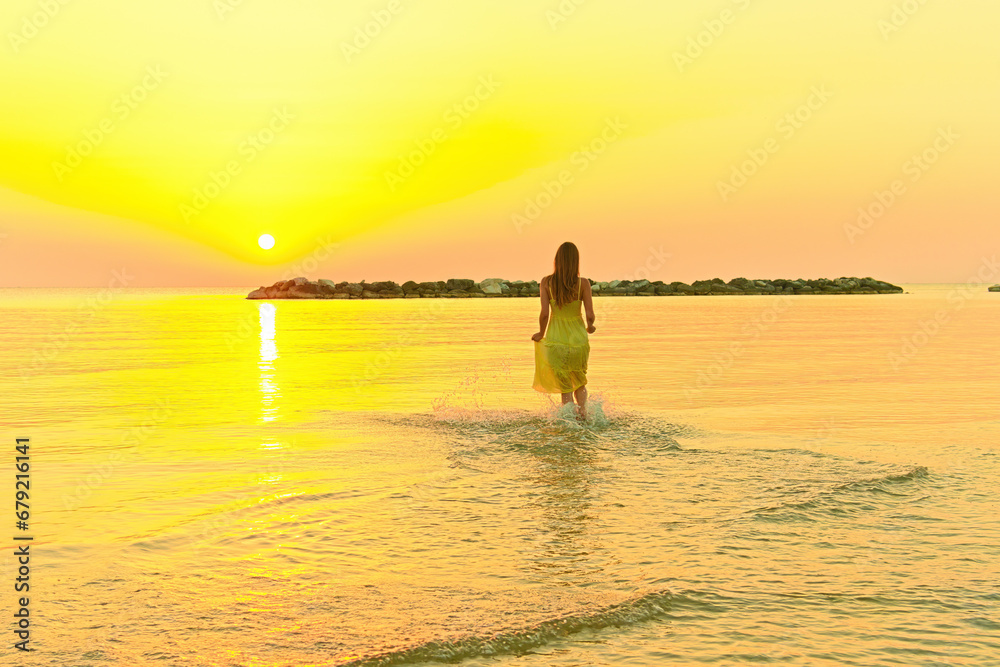A young woman in a bright yellow dress meets the sunrise, joyfully runs through the sea water.