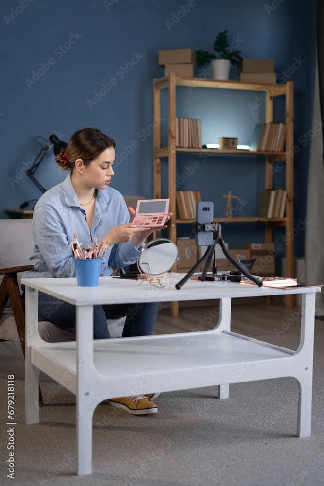 Beauty blogger woman filming daily make-up routine tutorial with eyeshadow palette on mobile phone. Influencer recording live streaming cosmetics product review in home studio