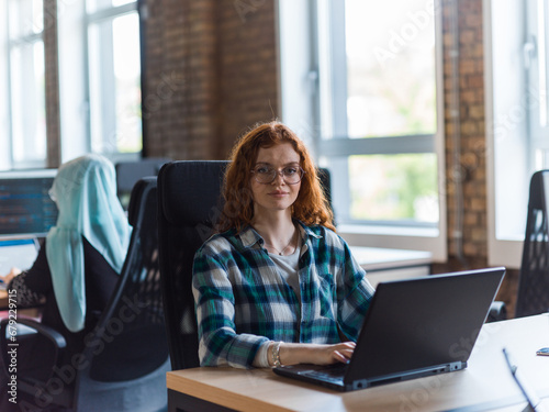 A young and successful businesswoman with vibrant orange hair engages in focused work within a modern office, showcasing her confidence and dynamic approach as she utilizes her laptop, emblematic of a