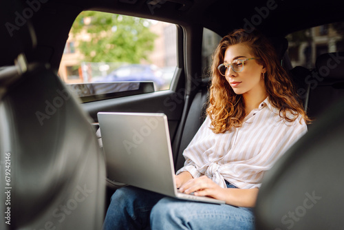 Beautiful business woman in casual clothes with a laptop working in the back seat of a car. Remote work concept, transport, technology. © maxbelchenko