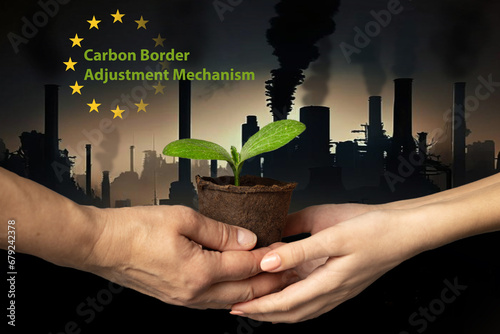 EU Carbon Border Adjustment Mechanism. Ecology. Hands of adult and child hold peat pot with green sprout of plant against backdrop of industrial cityscape with smoking chimneys and dark sky. CBAM.  photo