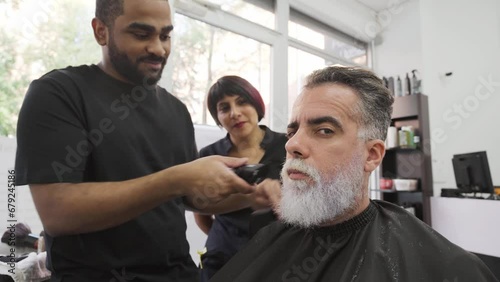 Client making faces and smirks at camera while stylists work on his beard.MOV photo