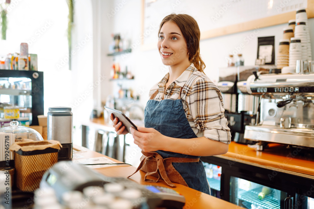 Portrait of a friendly female barista holding a tablet computer and smiling. A young coffee shop owner standing behind the bar using a digital tablet. Takeaway concept, technology. Portrait of a friendly female barista holding a tablet computer and smiling. A young coffee shop owner standing behind the bar using a digital tablet. Takeaway concept, technology.