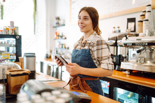 Portrait of a friendly female barista holding a tablet computer and smiling. A young coffee shop owner standing behind the bar using a digital tablet. Takeaway concept, technology.