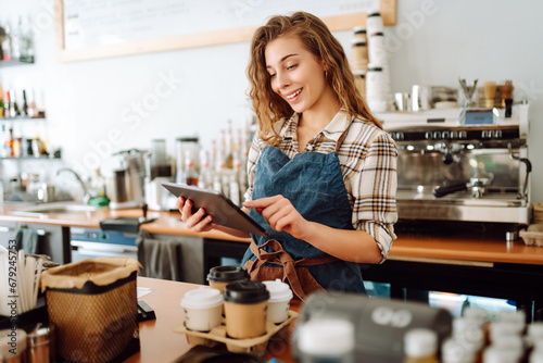 Portrait of a friendly female barista holding a tablet computer and smiling. A young coffee shop owner standing behind the bar using a digital tablet. Takeaway concept, technology.