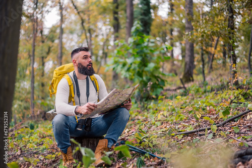 During his woodland hike, a hipster enjoys a rest, sitting on a tree stump while consulting a paper map to plan his route."