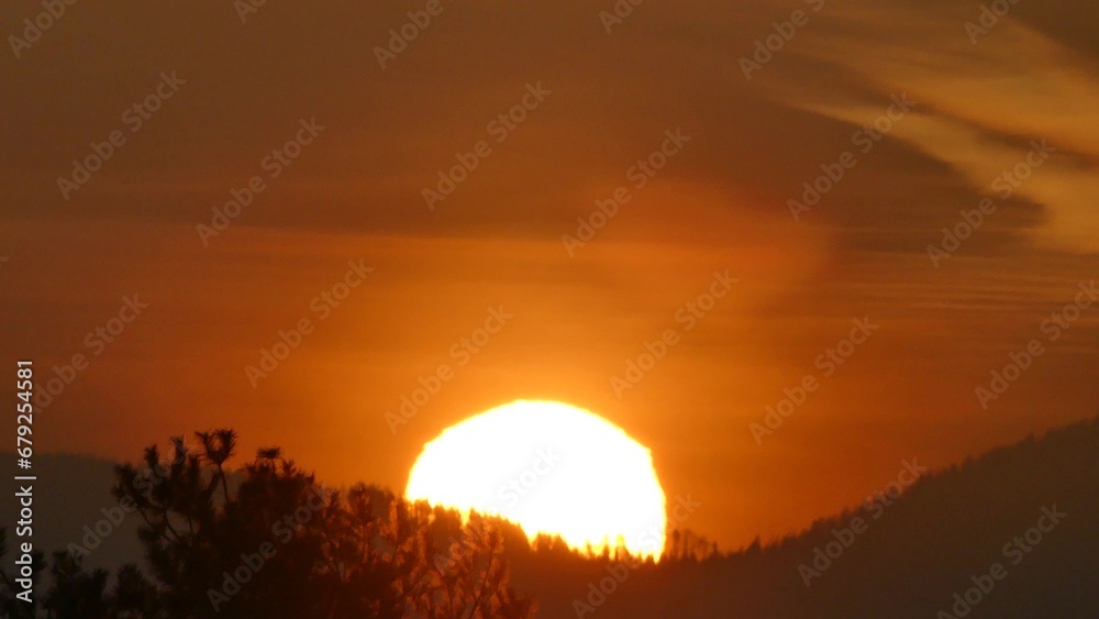 Orange sky and yellow sunrise over dark horizon of mountains with forests. Sun disc with Atmospheric refraction, distorted shape of sphere caused by atmospheric optical phenomenon.