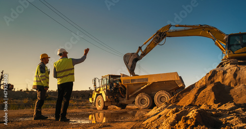 Caucasian Male Real Estate Investor And Civil Engineer Talking On Construction Site Of Apartment Block. Colleagues Discussing Building Progress. Excavator Loading Sand In Industrial Truck On Warm Day photo
