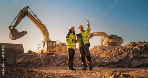 Hispanic Female Inspector Talking to Caucasian Male Land Development Manager With Tablet On Construction Site Of Real Estate Project. Excavators Preparing For Laying Building Foundation. Hot Sunny Day photo