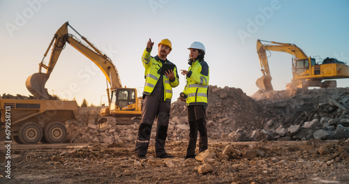 Caucasian Male Civil Engineer Talking To Hispanic Female Inspector And Using Tablet On Construction Site of Apartment Complex. Real Estate Developers Discussing Business, Excavators Working.