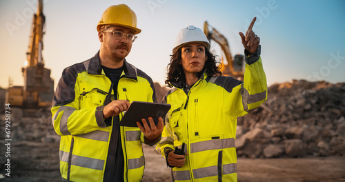 Caucasian Male Civil Engineer Talking To Hispanic Female Inspector And Using Tablet Computer On Construction Site of New Building. Real Estate Developers Discussing Business, Excavators Working.