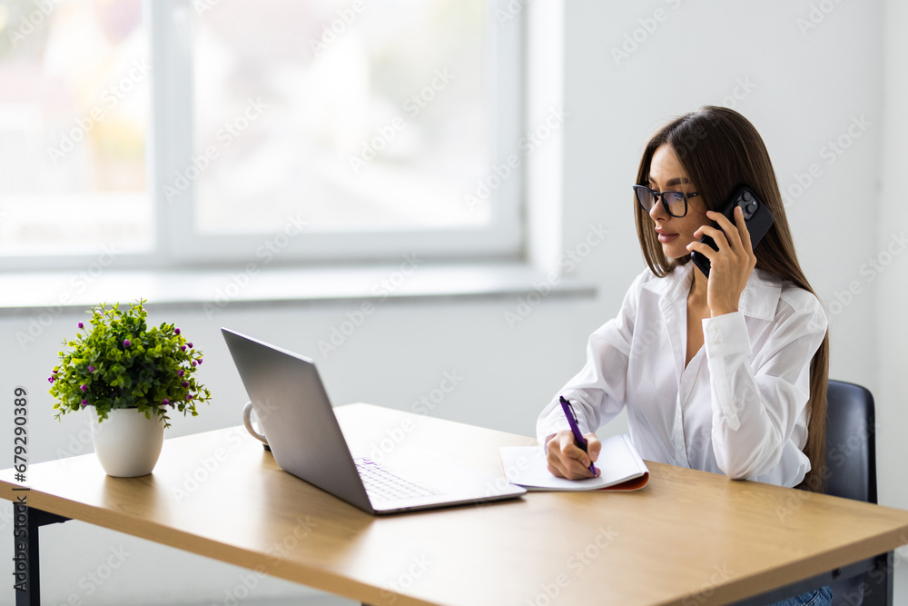 Young woman sales manager talking on cell phone using laptop in office Young woman sales manager talking on cell phone using laptop in office