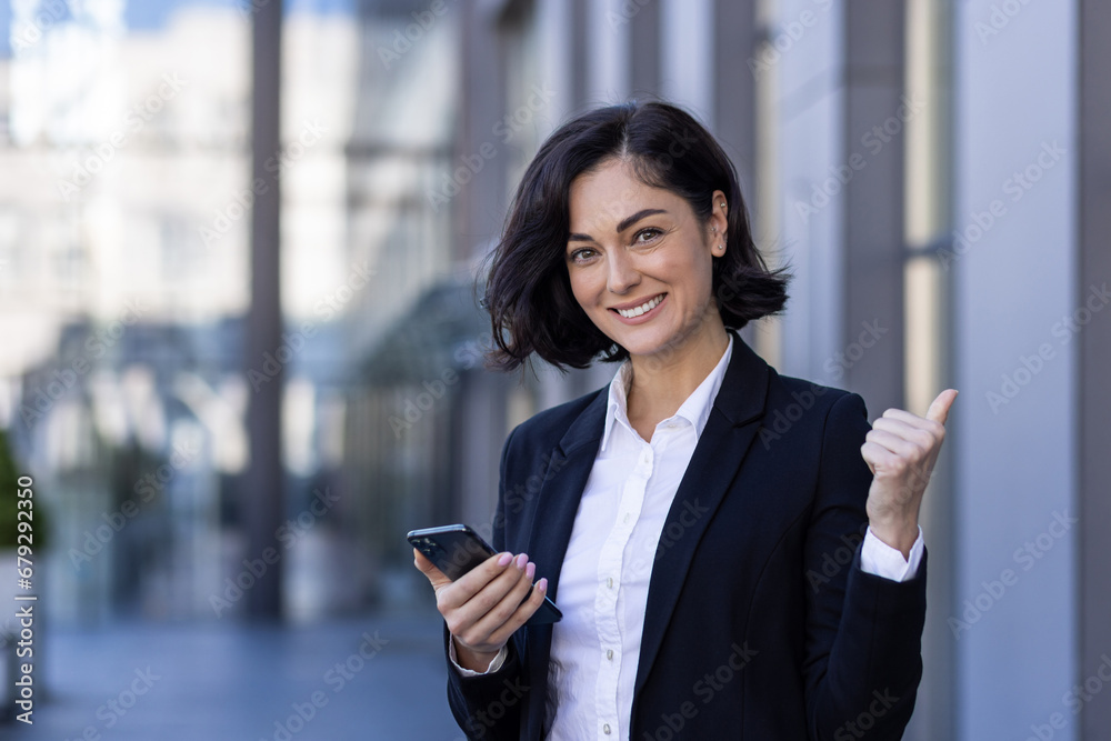 Portrait of a young successful business woman standing near an office center, holding a phone, showing a super finger and smiling at the camera