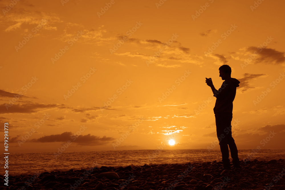 Fototapeta premium black silhouette of Man taking a photo with his mobile phone during the lovely sunset in Black sea. Male traveler make panorama shoot. adult man standing on empty yellow orange beach on background.