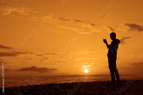 black silhouette of Man taking a photo with his mobile phone during the lovely sunset in Black sea. Male traveler make panorama shoot. adult man standing on empty yellow orange beach on background.