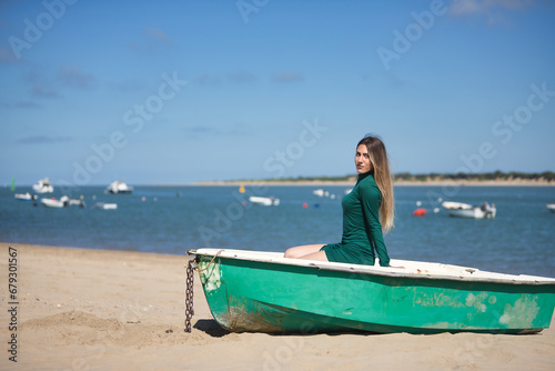 Young, pretty, blonde, green-eyed young woman in a green dress, sitting in a boat, looking at the camera, relaxed and calm, on the beach, with the sea in the background. Concept vacation, sun, travel