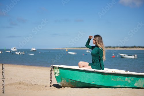 Young, beautiful, blonde, green-eyed, young woman in a green dress, sitting in a boat, covering herself from the sun with her hand, relaxed and calm, on the beach, with the sea in the background. © Manuel